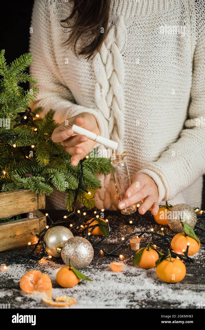 girl in knitted sweater makes a Christmas and puts it in little glass bottle, mandarines and fir tree decoration behind Stock Photo