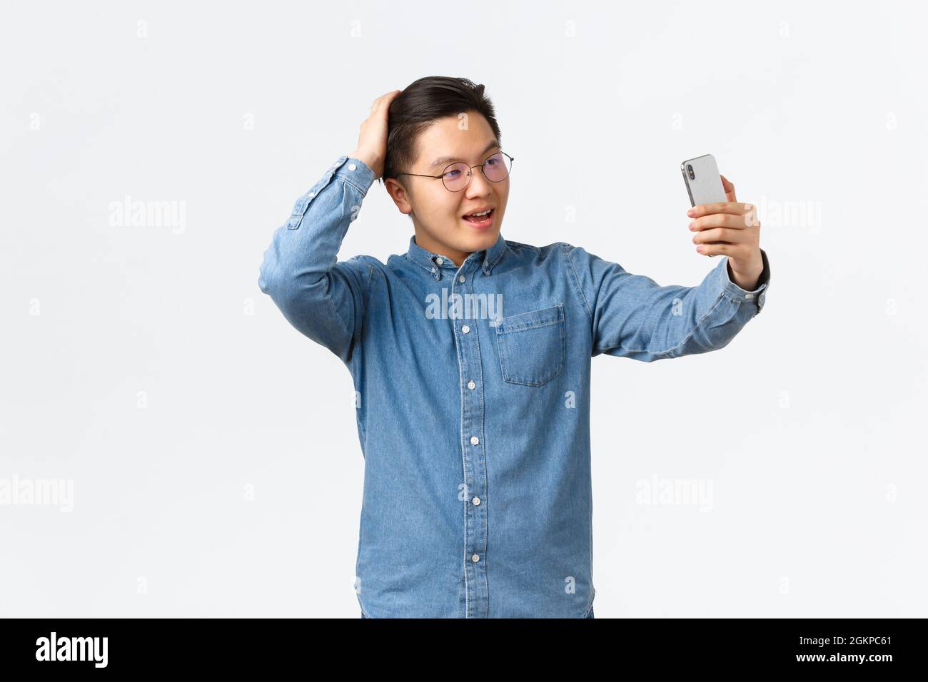 Cheeky confident asian man in glasses and braces feeling sassy, taking selfie, brushing hair with hand, posing for photo, using filter app on mobile Stock Photo