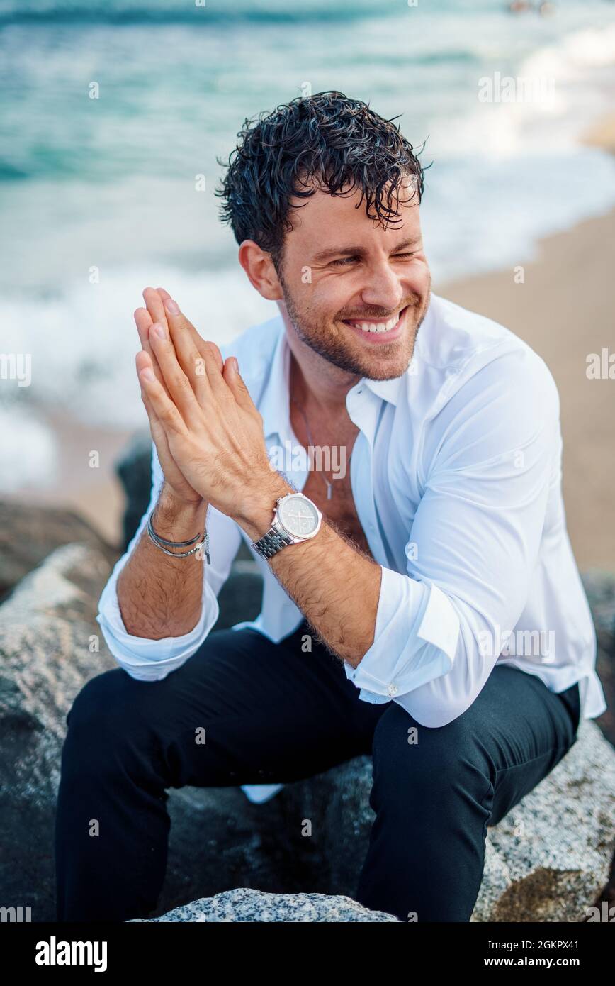 Happy masculine male in stylish white shirt and with wet hair sitting on rock at seaside and looking away Stock Photo