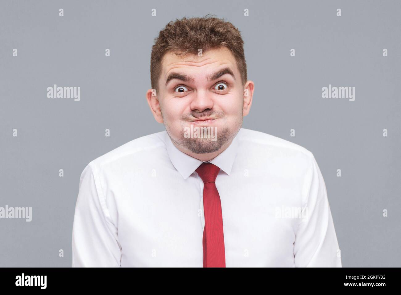 funny portrait of young man in white shirt and tie standing blowing his cheeks and looking at camera with funny face and big eyes. indoor isolated on Stock Photo