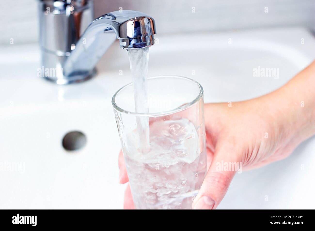 Young caucasian woman hand holding a glass with pure drinking water pouring from home faucet close up. Stock Photo