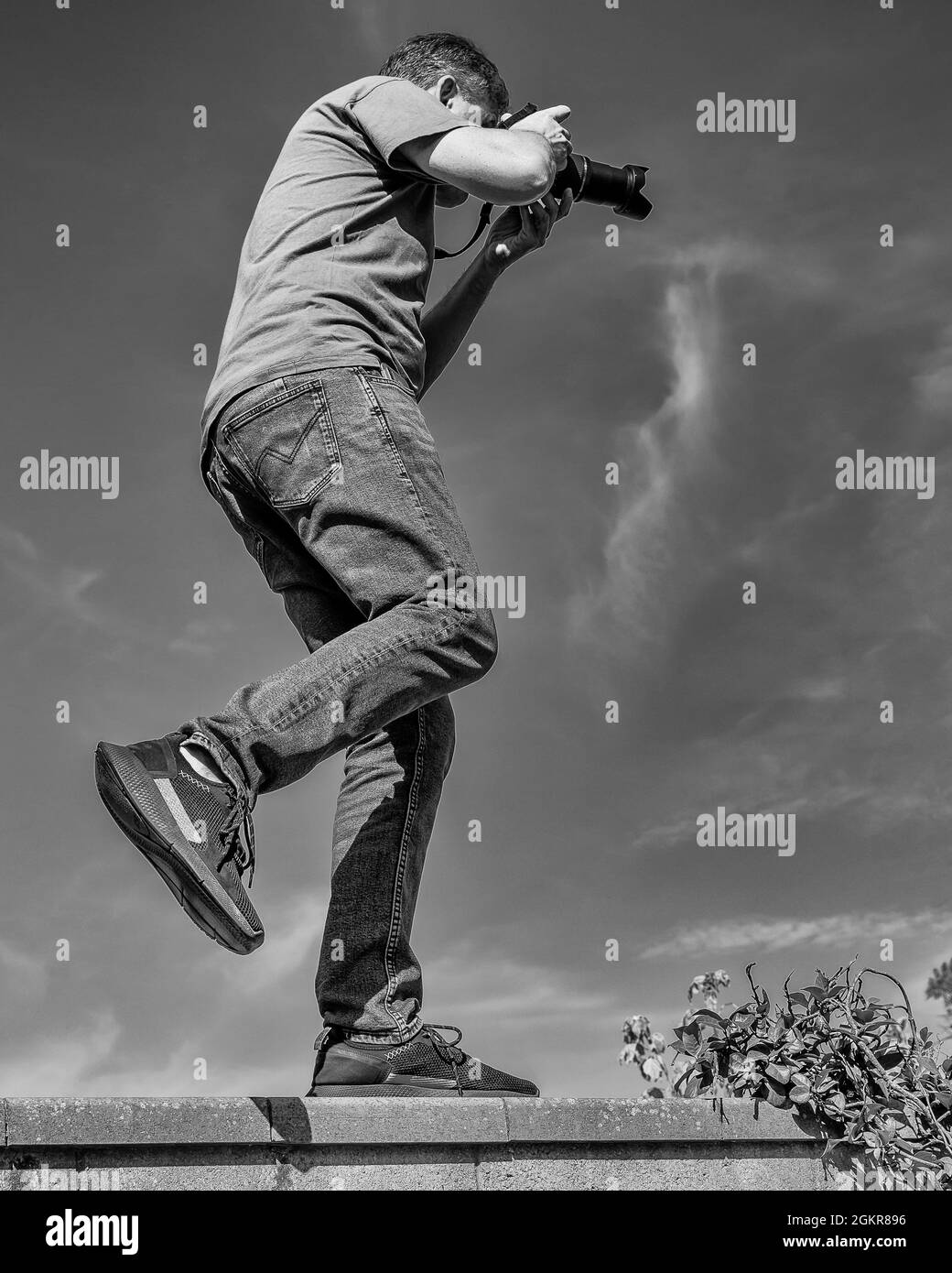A photographer balances precariously on one leg as he takes a photo from above a narrow wall, in black and white Stock Photo