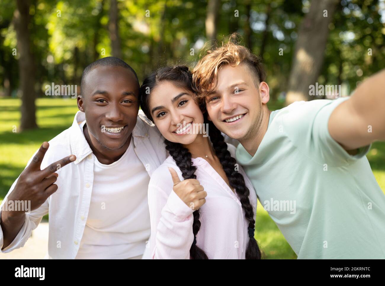 Multiracial students friends making selfie, gesturing v-sign and showing thumb up, posing outdoors in campus Stock Photo