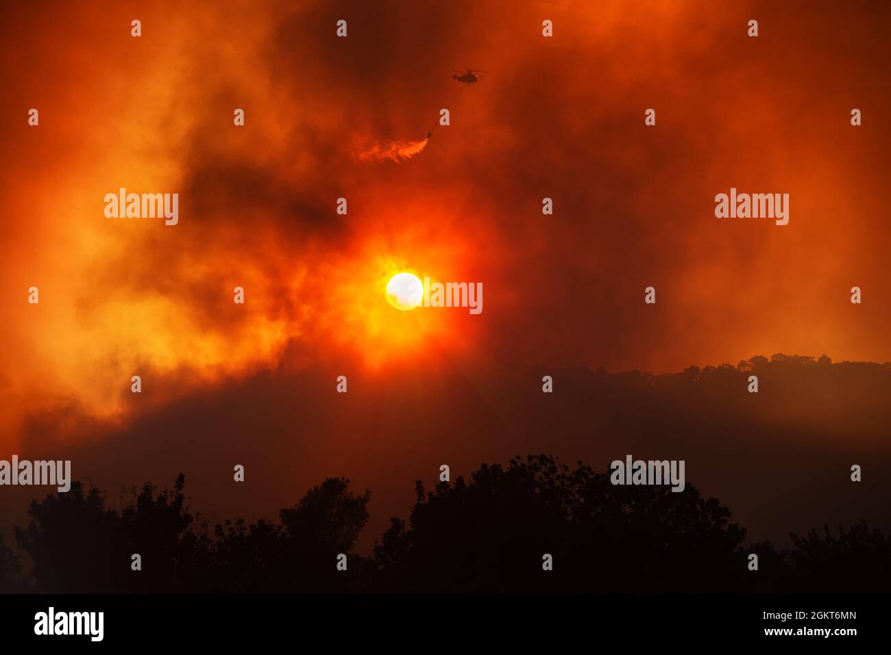 Helicopter dropping water from the bucket, fighting forest fire. Dramatic landscape against the setting sun with red sky and heavy smoke Stock Photo