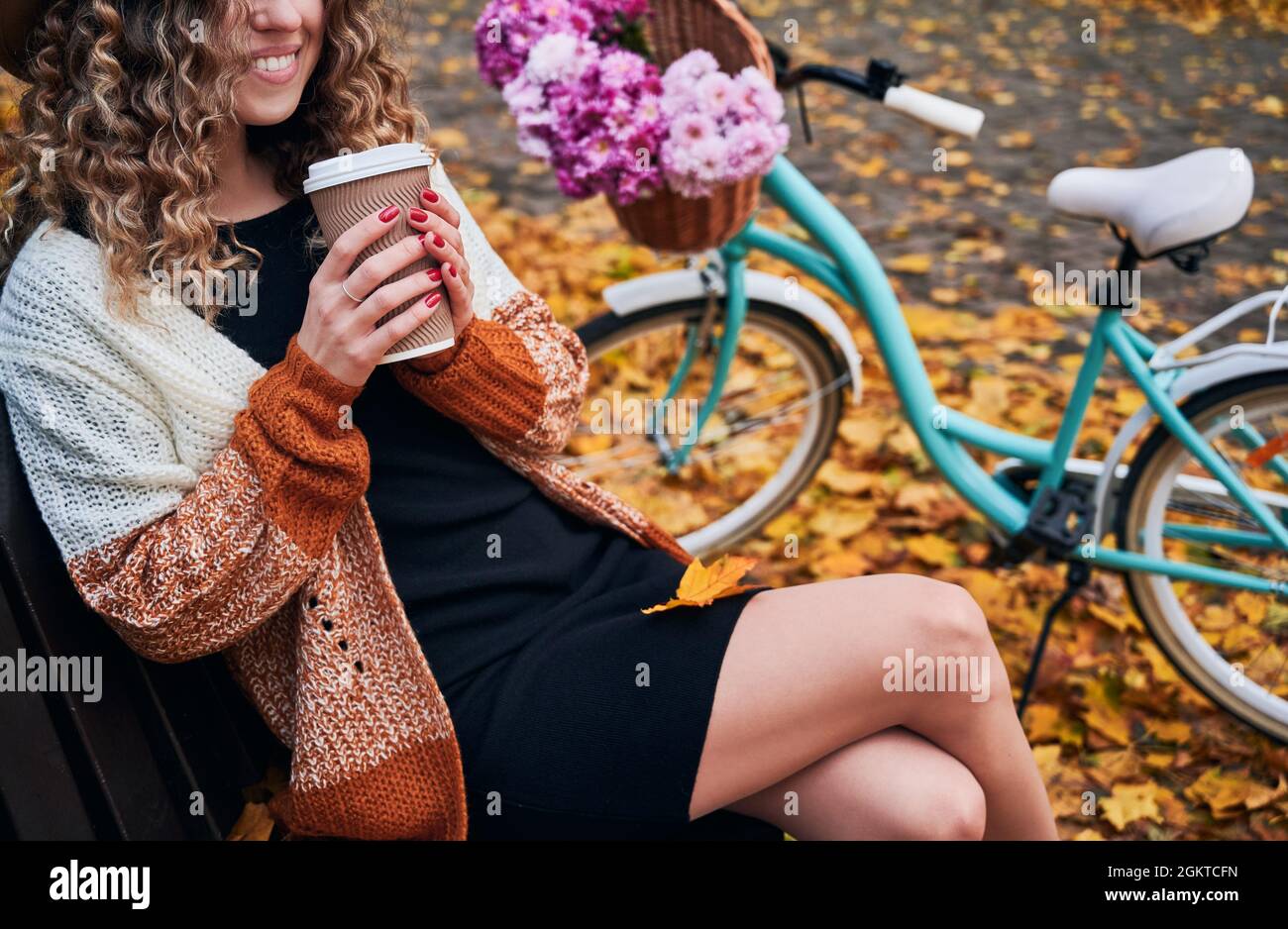 Smiling young woman sitting on bench near bike with flowers and drinking coffee to go in autumn park. Elegant woman holding cup of hot drink and smiling while resting outdoors near city bicycle. Stock Photo