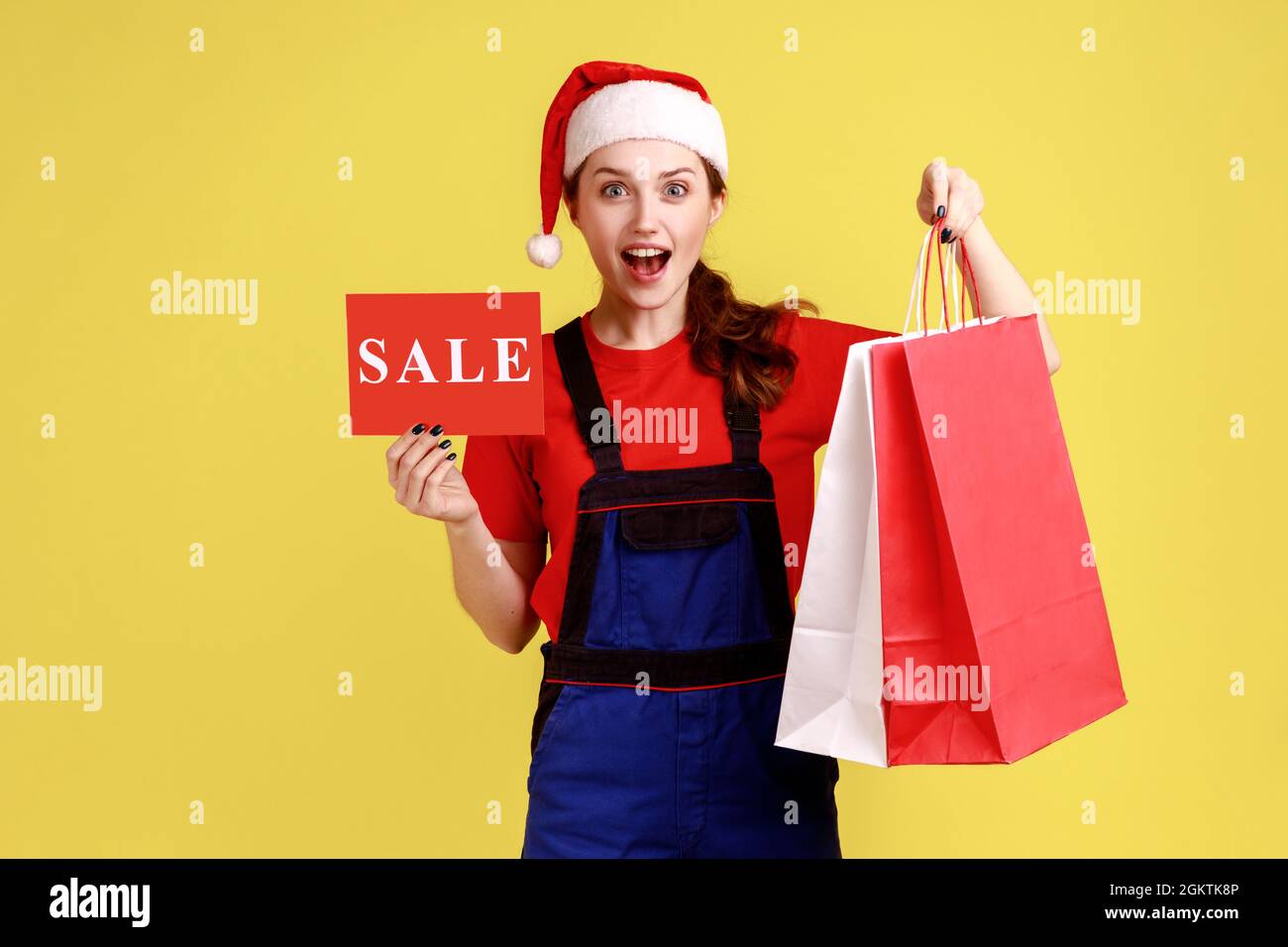 Surprised courier female with sale card and shopping bags, presents discounts for delivering orders, wearing blue overalls and santa claus hat. Indoor Stock Photo