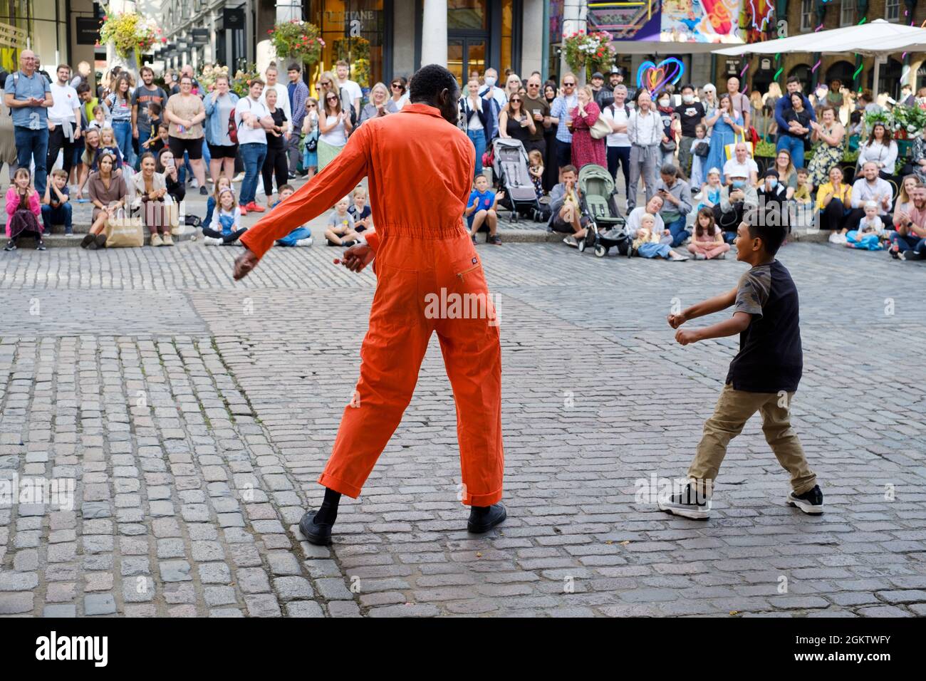 A street entertainer or street performer performs with a child, selected from the watching crowd, and both dance the floss Stock Photo