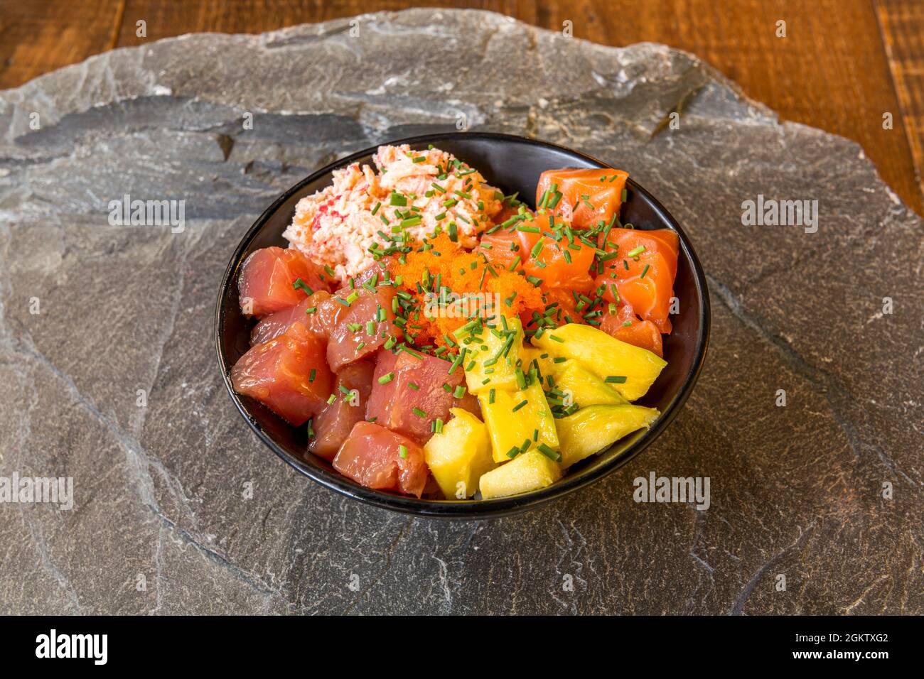 Mixed Hawaiian poke bowl with salmon, fish roe, red tuna with mango and surimi and lots of chopped chives on a gray stone background. Stock Photo