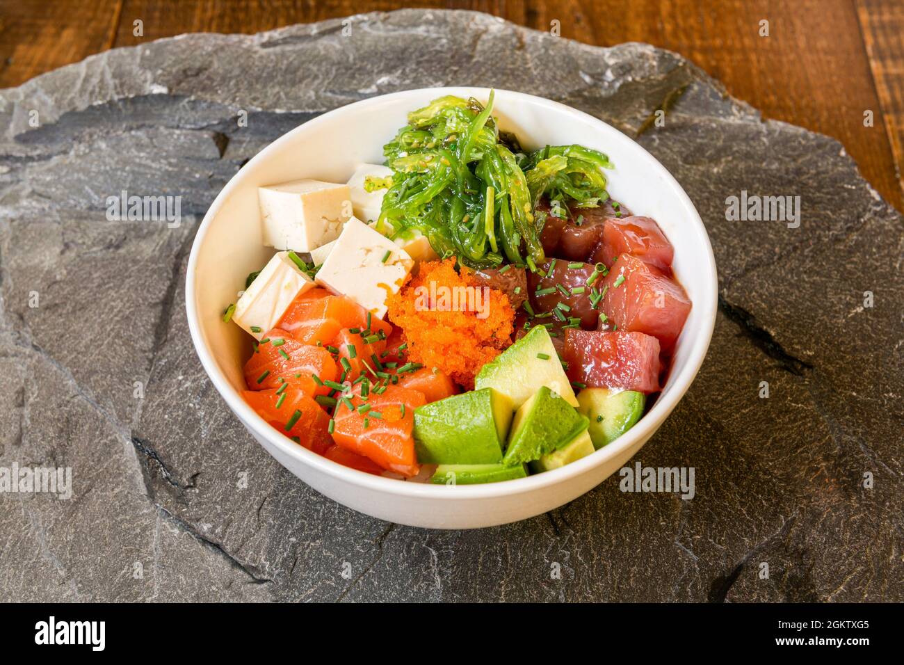 Poke bowl with fish roe, salmon, red tuna, green avocado, white tofu and guacamole seaweed with sesame seeds on gray stone background Stock Photo