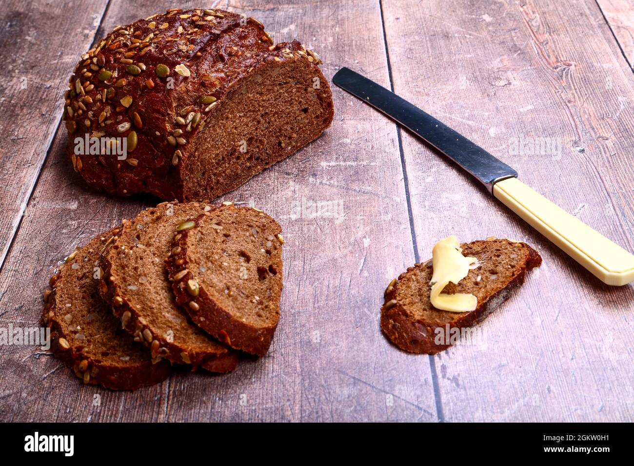 Freshly baked Pumpernickel Boule artisan bread with buttered bread slices cut and laid on a wooden table with a old bred knife Stock Photo