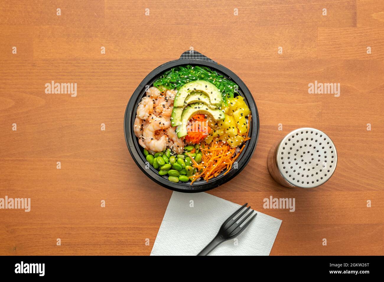 Cooked prawn poke bowl with edamame beans, grated carrot, diced mango, avocado slices, orange fish roe and wakame seaweed salad in a black home delive Stock Photo