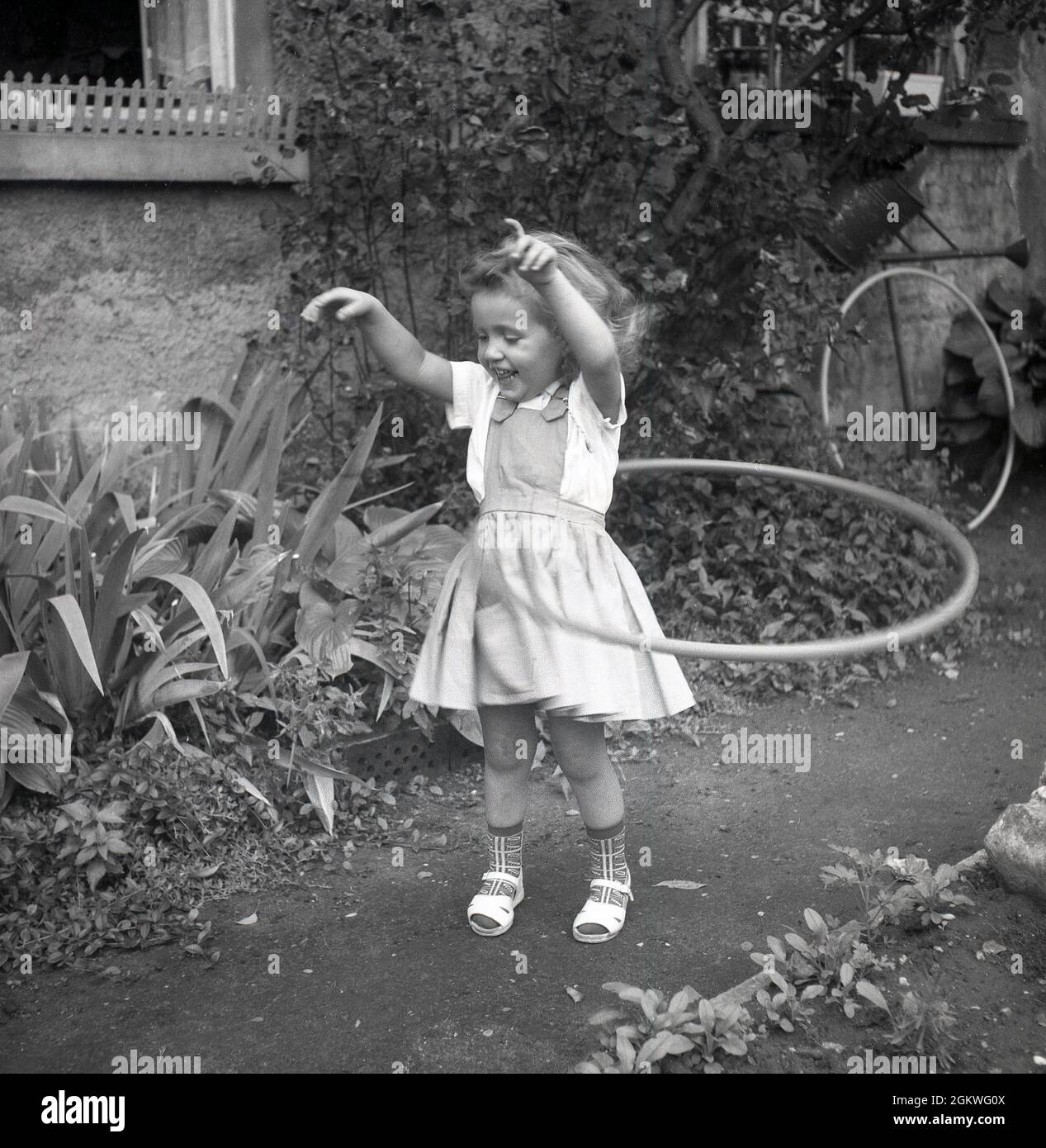 1960s, historical, outside on a garden path, a little girl playing with a hula hoop, a toy hoop that she is twirling around her waist, Germany. Although a hoop had been in existence and used by children for hundreds of years - but not known by that name - it was only in the late 1950s, when the plastic version was introduced, that the craze for them took off. The hula hoop - the name coming it is said from doing the Hula, the dance in Hawaii - became a world-wide fad, with more than 100 million sold in two years. Hula hoops remained a popular children's toy until the late 1970s, early 80s. Stock Photo