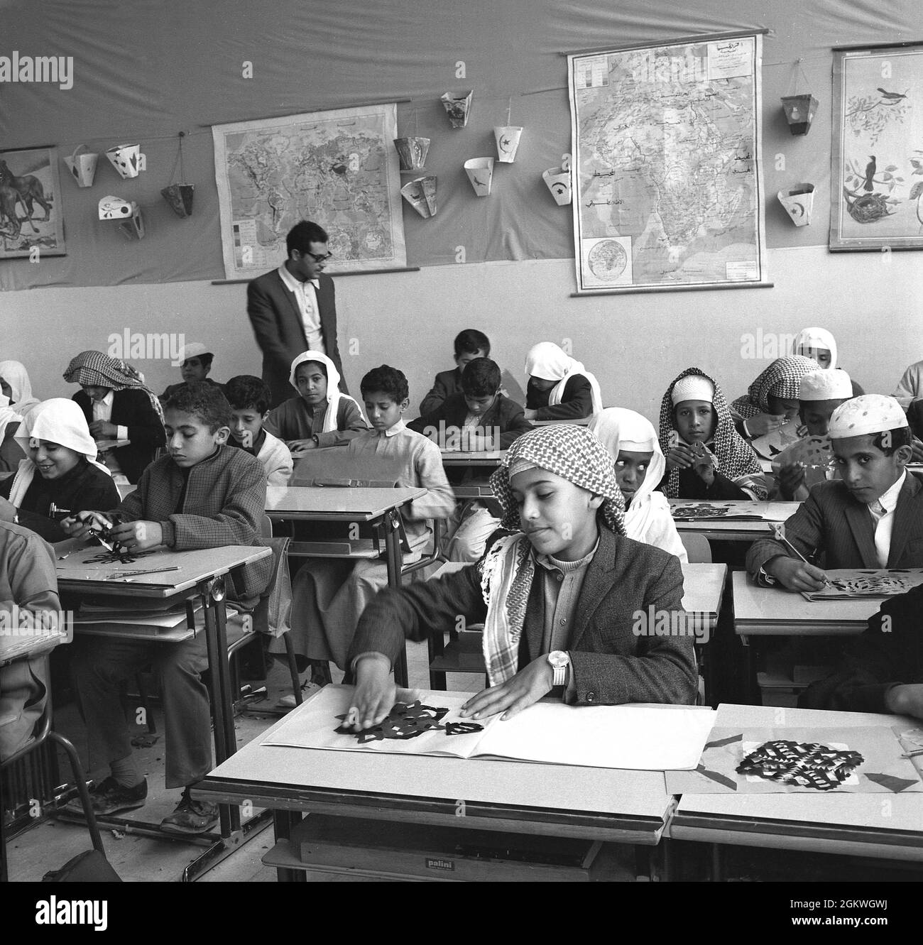 1960s, historical, young arab schoolboys, some with headdresses on, sitting in a classroom at a primary school, with male teacher, cutting and making shapes and patterns from card. Stock Photo