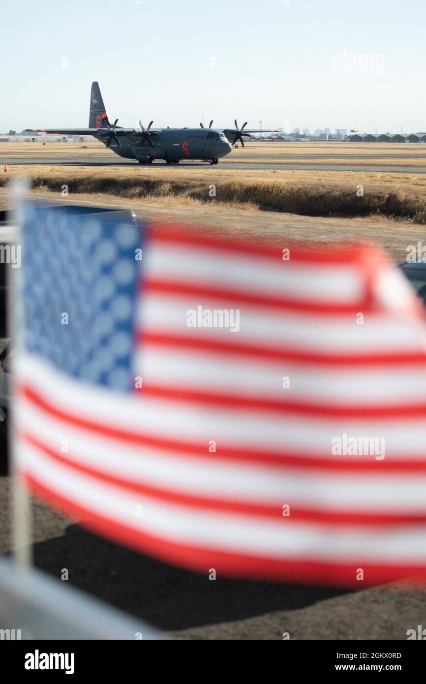 A California Air National Guard C-130 taxis to the runway July 14, 2021 at CAL FIRE Air Tanker Base, McClellan Park, Calif. as the U.S. Flag waves in the wind. The Air Force C-130 MAFFS-equipped aircraft, out of Channel Islands, Calif. was requested by the National Interagency Fire Center and approved by the Secretary of Defense, are providing unique fire-fighting capabilities. The Air Force MAFFS teams are managed by First Air Force (Air Forces Northern)’s 153rd Air Expeditionary Group. Stock Photo