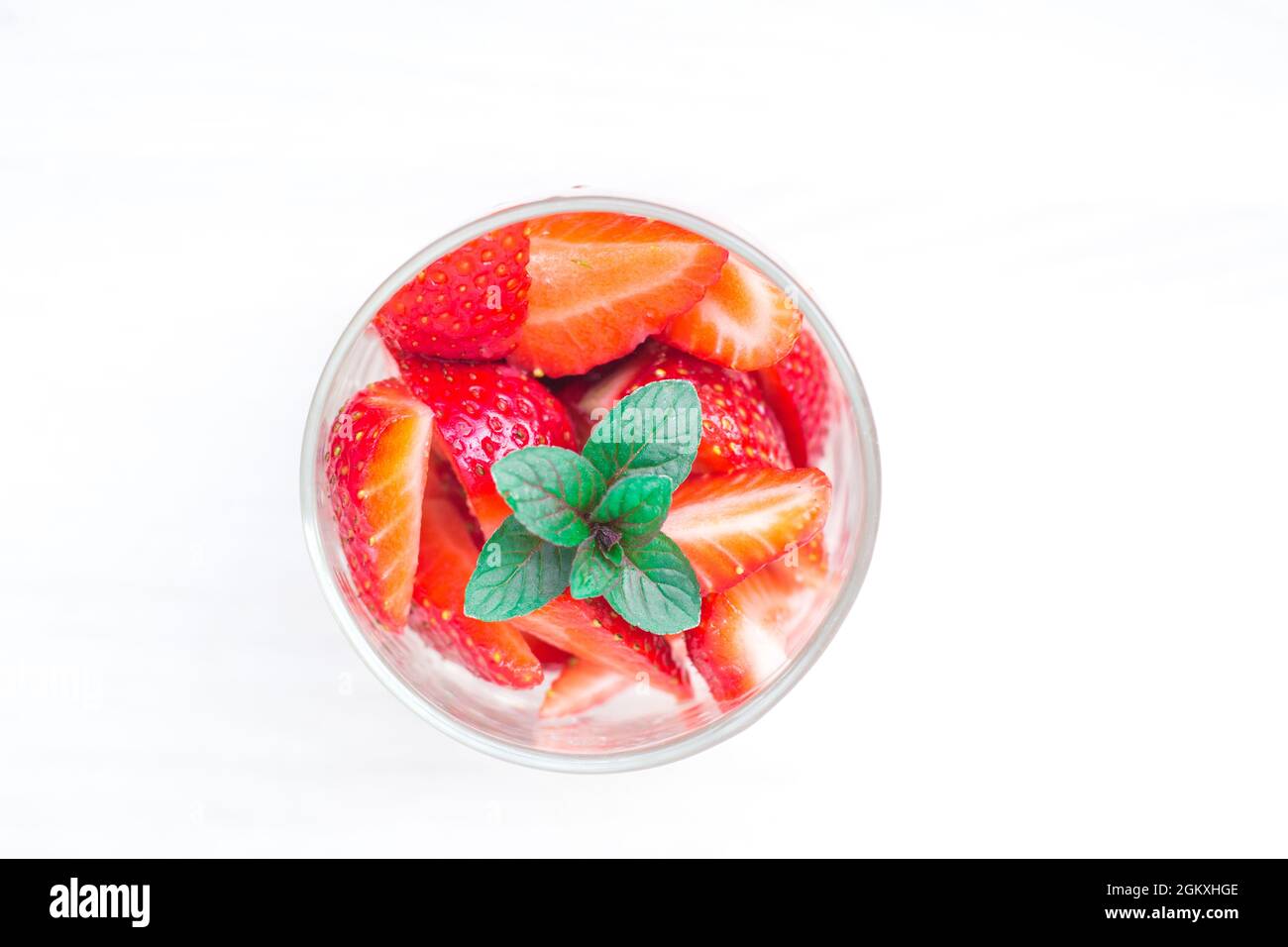 Glass with pieces of red strawberry and fresh peppermint leaves on top. Top view to a round dessert on white wooden background. Stock Photo