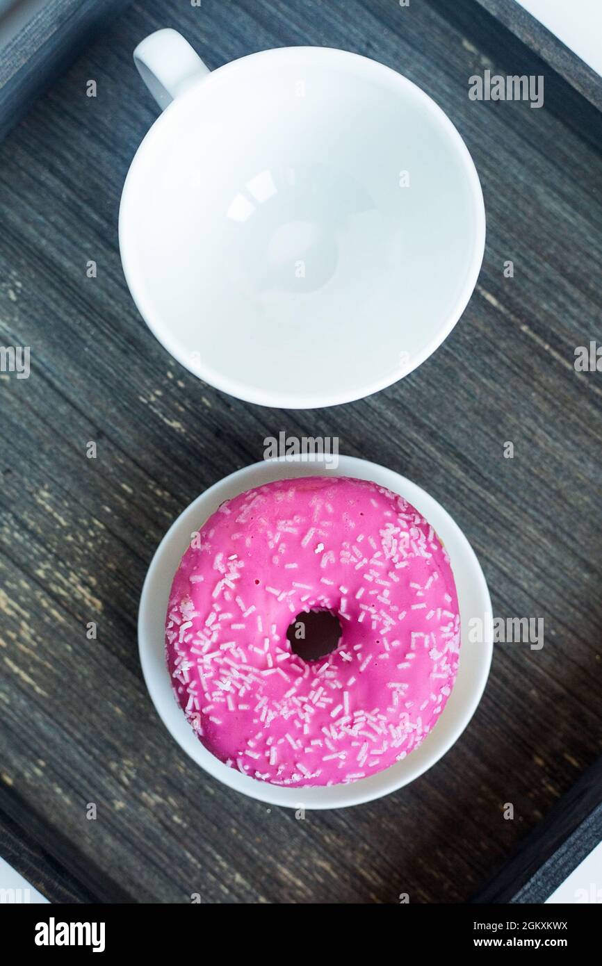 Vertical picture of bright pink donut on white round plate and tea cup above. Aged wooden background. Isolated objects. Traditional American breakfast Stock Photo