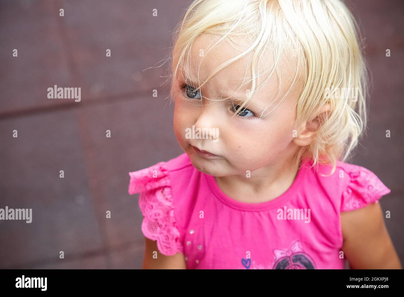 Serious angry baby girl looking left. Blond blu eye kid in pink t-shirt portrait. Blurred protective rubber pavement of playground in background Stock Photo