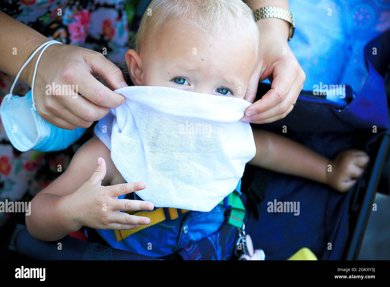 Mother puts white scarf as a protective musk on blue eye blond baby. kids portrait. Female caucasian hand with blue medical mask on wrist. Looking Stock Photo