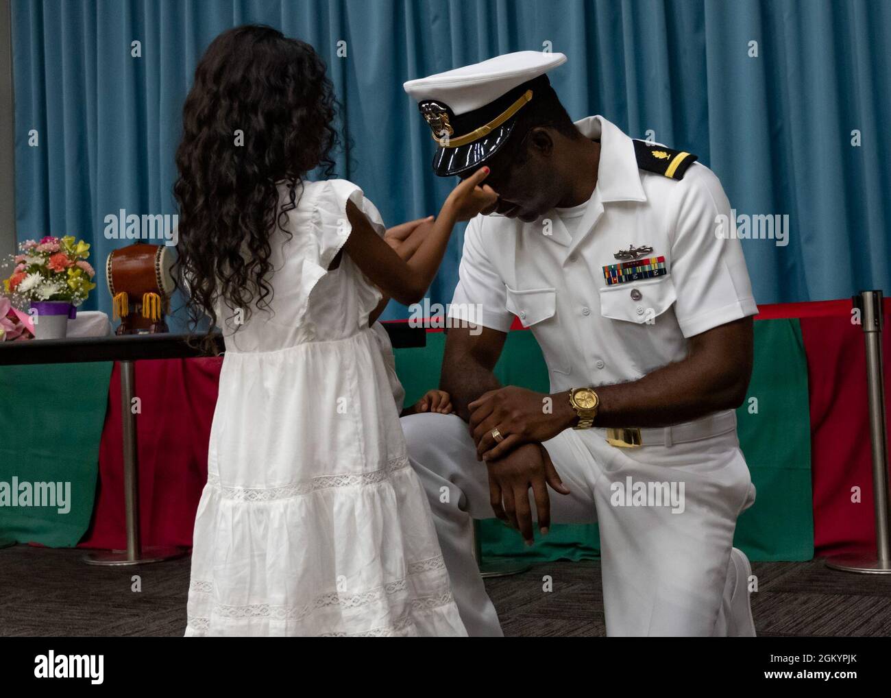 CAMP SHIELDS, Japan (July 30, 2021) Ensign Frederick Joshua, from Baltimore, MD and assigned to Commander, Fleet Activities Okinawa security department, kneels so his daughter may help him don his commissioned officer’s combination cover during his formal commissioning ceremony at Camp Shields, Okinawa, Japan July 30, 2021. Joshua was commissioned into the U.S. Naval Medical Service Corps (MSC) through the MSC In-Service Procurement Program. It allows Sailors in pay grades E-5 through E-9 who meet certain eligibility criteria to directly commission into MSC and attend fully-funded training to Stock Photo