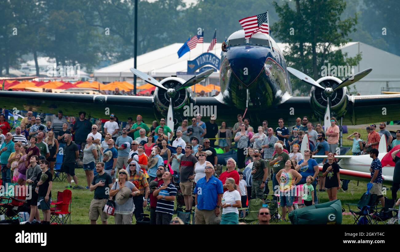 Airshow attendees stand for the national anthem during a gunship legacy flight as part of EAA AirVenture Oshkosh 2021, at Wittman Regional Airport, Wis., July 31, 2021. Air Force Special Operations Command was featured at EAA’s airshow and brought multiple aircraft in its inventory to include the AC-130J Ghostrider, MC-130J Commando II, EC-130J Commando Solo, CV-22 Osprey, C-145 Combat Coyote, U-28 Draco, C-146 Wolfhound, MC-12 Liberty,  and the MQ-9 Reaper displaying capabilities of airpower through aerial demonstrations and/or static displays. Stock Photo