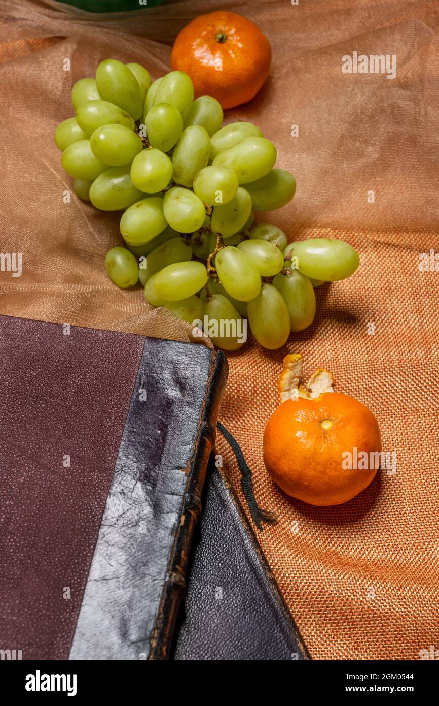 Bunch of elongated white dessert grapes with ripe tangerines on orange cloth and old books Stock Photo