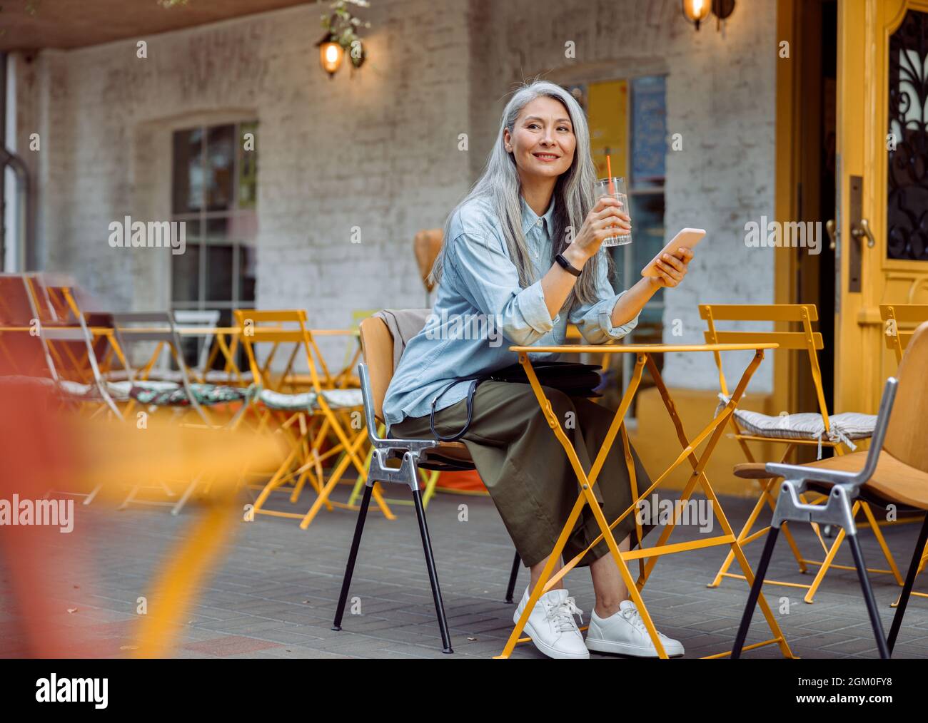 Grey haired Asian woman with glass and smartphone on outdoors cafe terrace Stock Photo