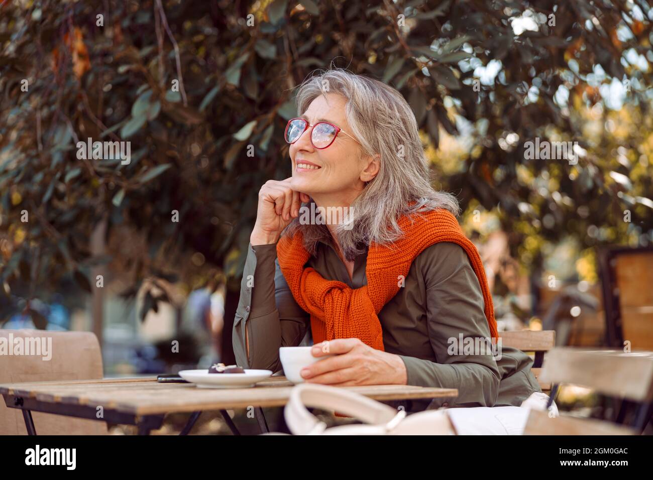 Dreamful mature lady with glasses sits leaning on hand at table on outdoors cafe terrace Stock Photo