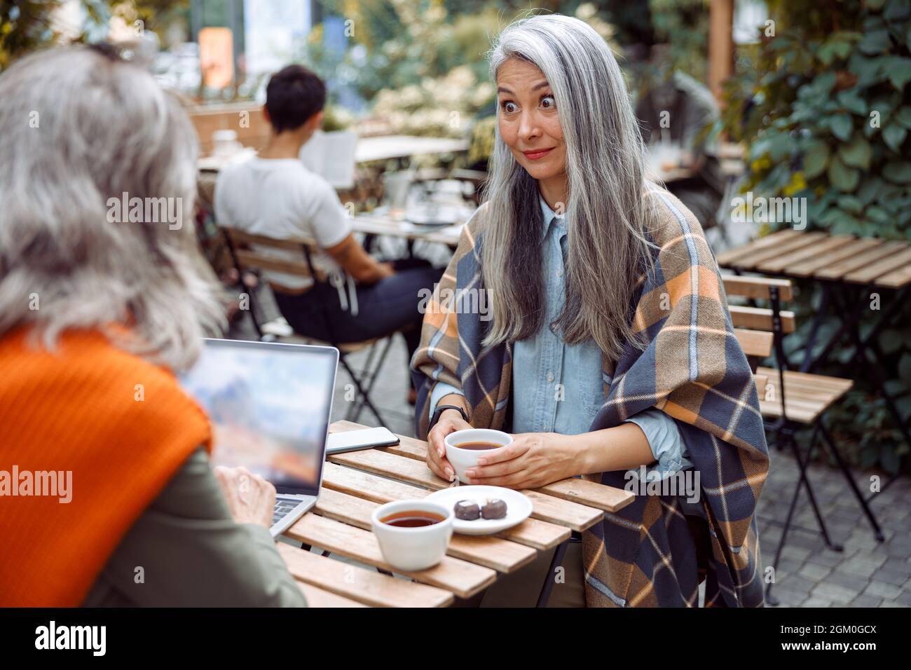 Surprised mature Asian lady looks at friend using laptop at small table in street cafe Stock Photo