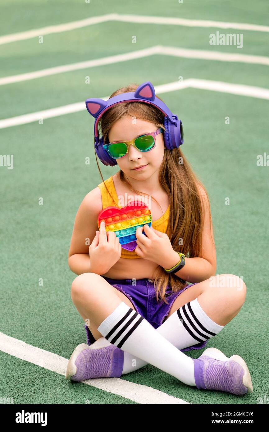 Girl play with popular pop It fidget toy. Child relaxing and chilling on sports ground feel no stress free. Preteen schoolgirl with wireless headphones listening to music. . Stock Photo