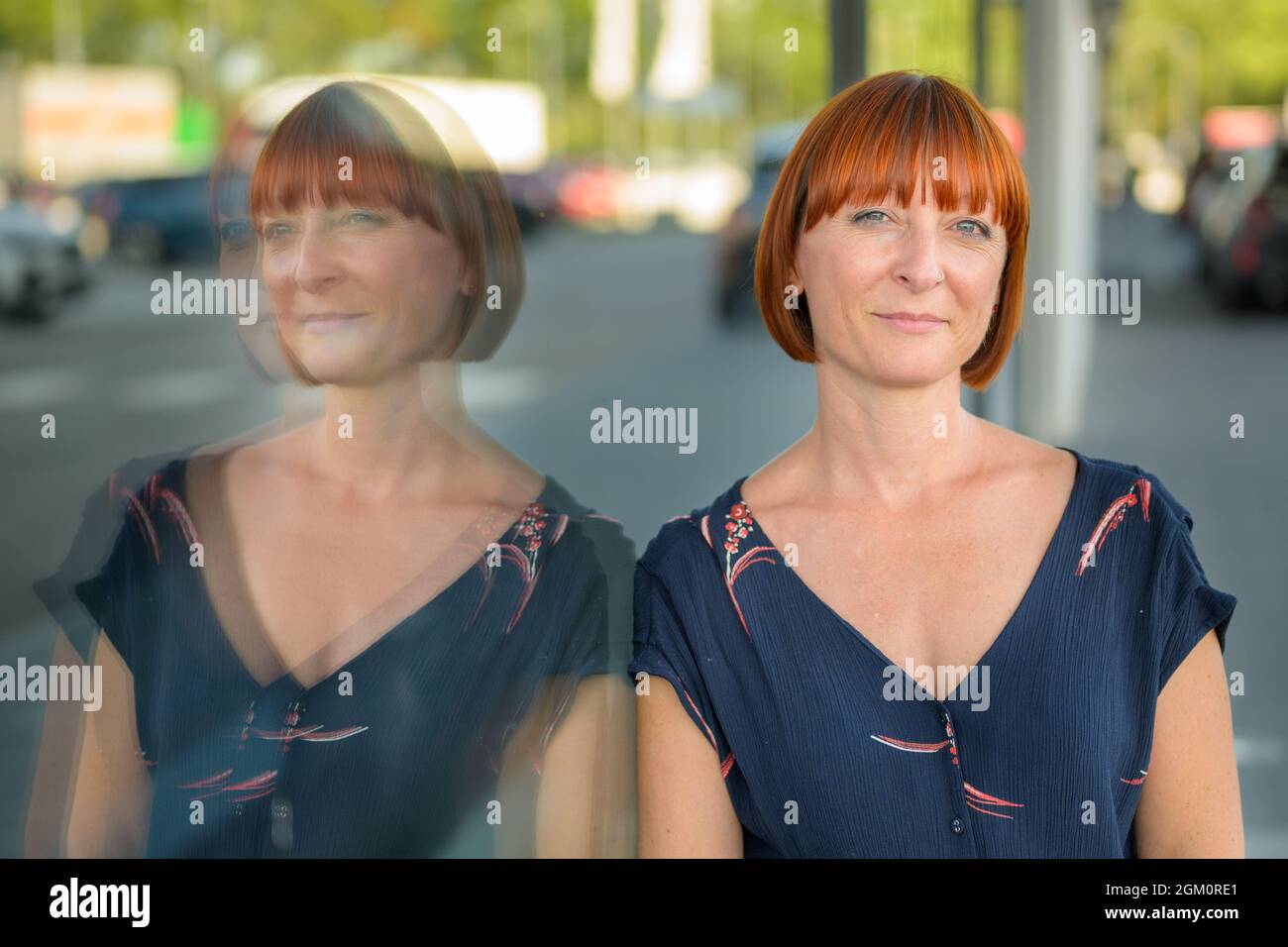 Thoughtful enigmatic woman looking quietly at the camera with a smile as she leans against a large window reflected in the glass in an urban street in Stock Photo