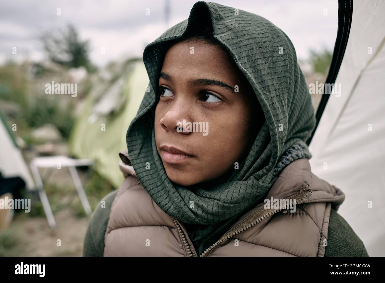 Sad Black refugee girl with covered head looking up hopefully, tent camp for migrant behind her Stock Photo