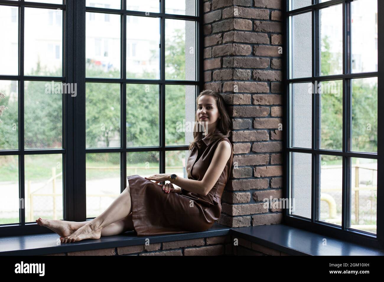 Beautiful young woman in brown leather dress sits on the windowsill ...