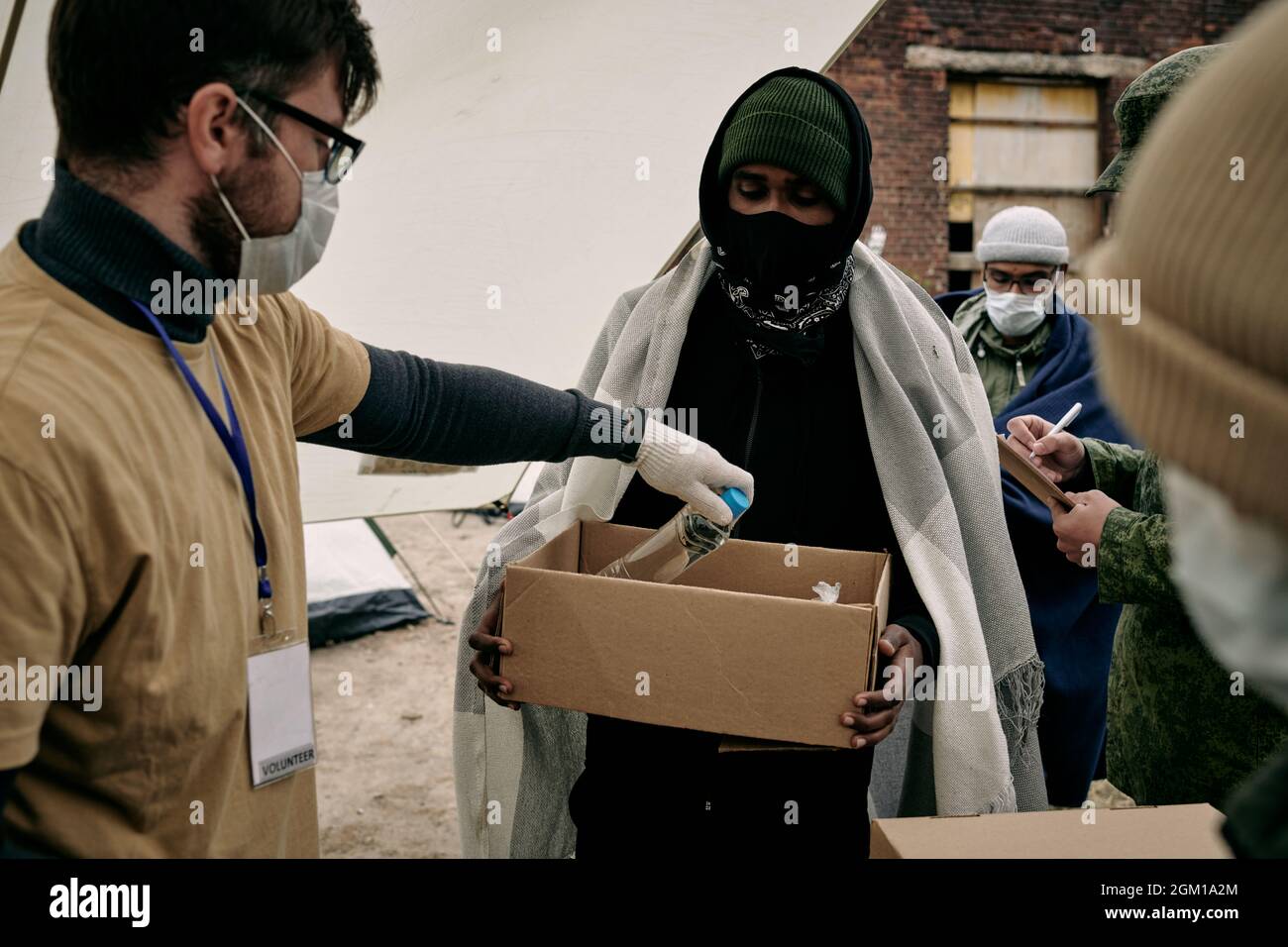 Social Worker in mask and glasses putting bottle of water into box held by middle-eastern refugee with plaid on shoulders Stock Photo