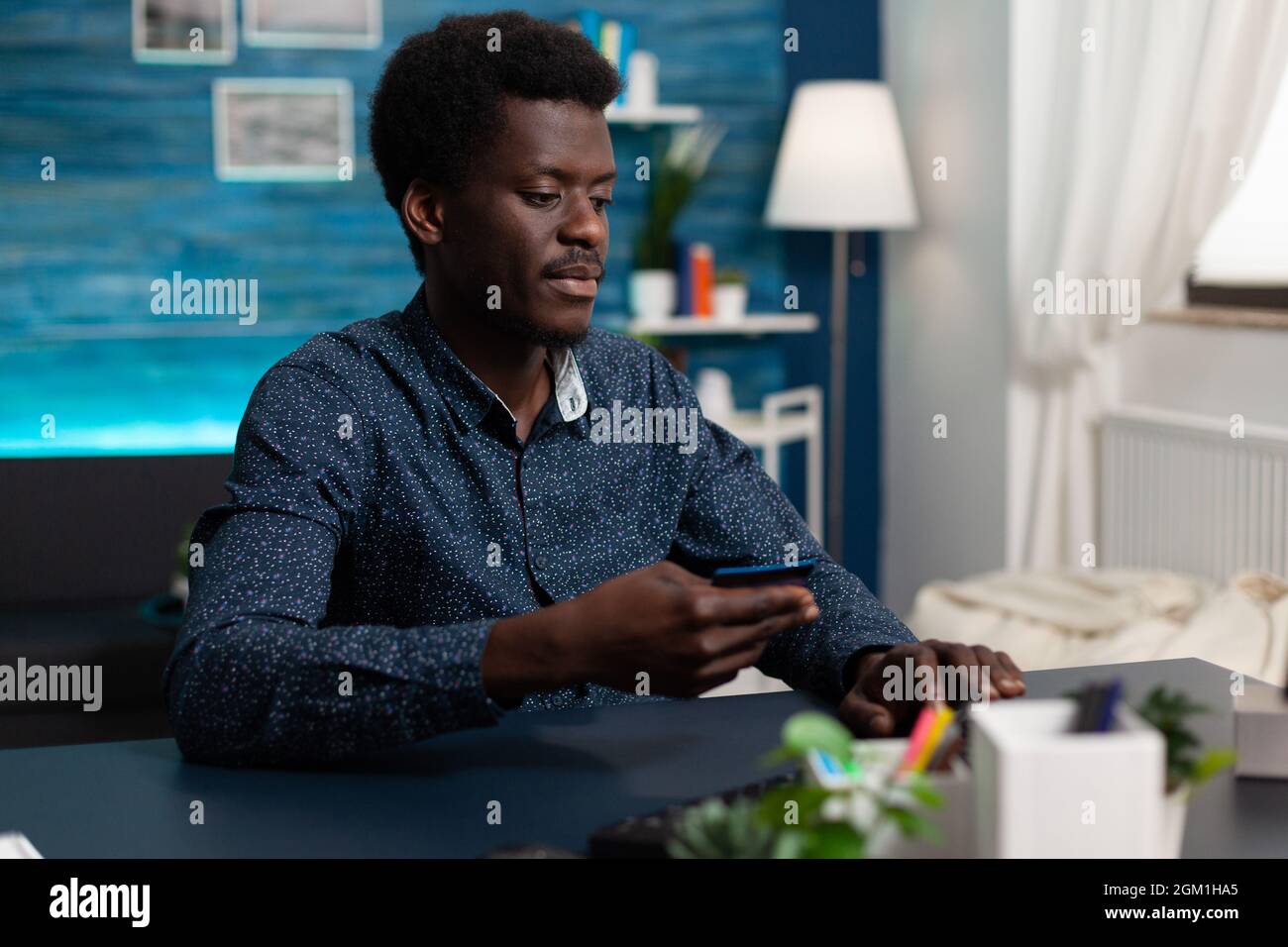 African consumer making online shopping on sale store paying order with credit card. Student sitting at desk in living room doing online payment with electronic wallet using computer Stock Photo