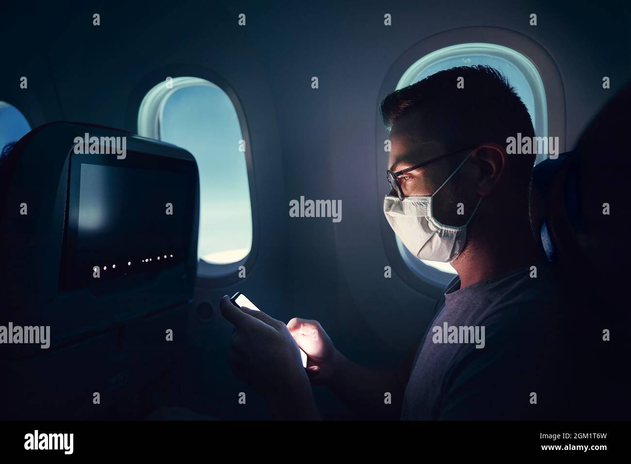 Man with face mask traveling by airplane. Passenger using phone during flight. Stock Photo