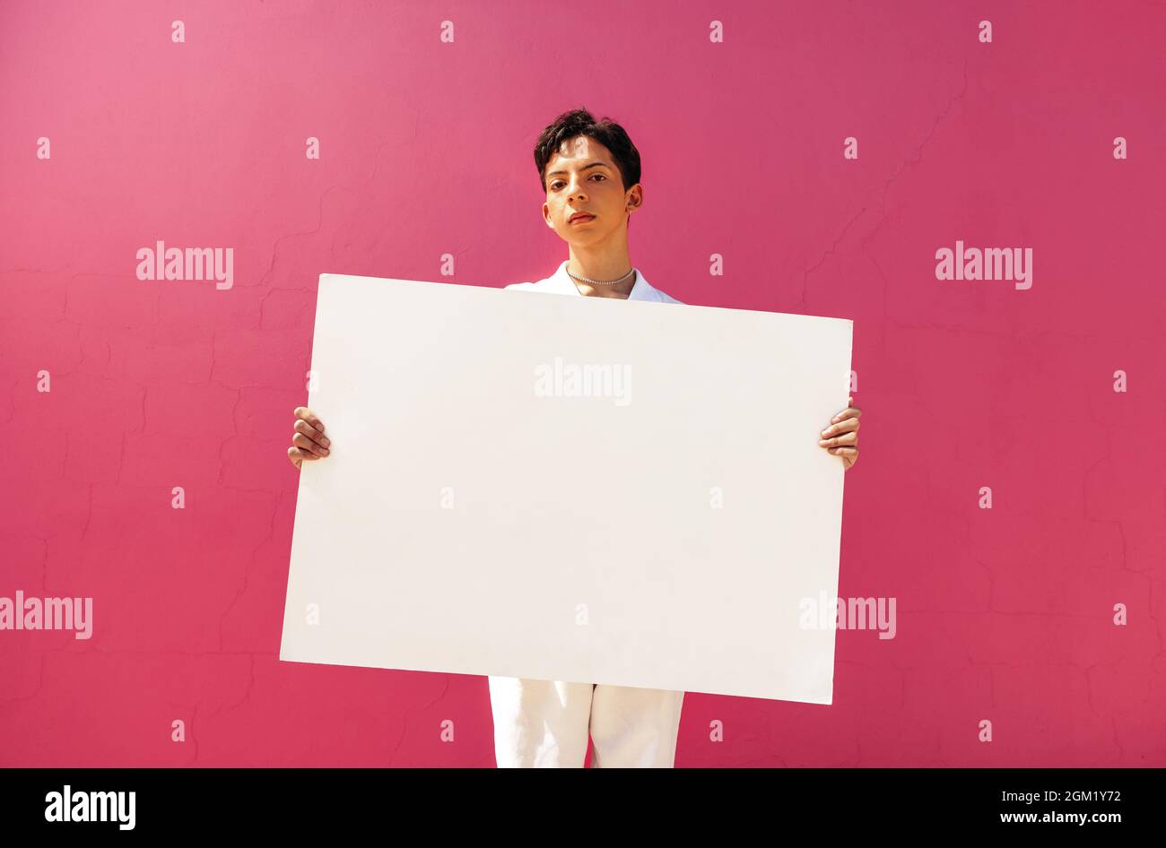 Confident queer boy holding a blank placard against a pink background. Assertive teenage activist displaying a white banner in a studio. Young gay boy Stock Photo