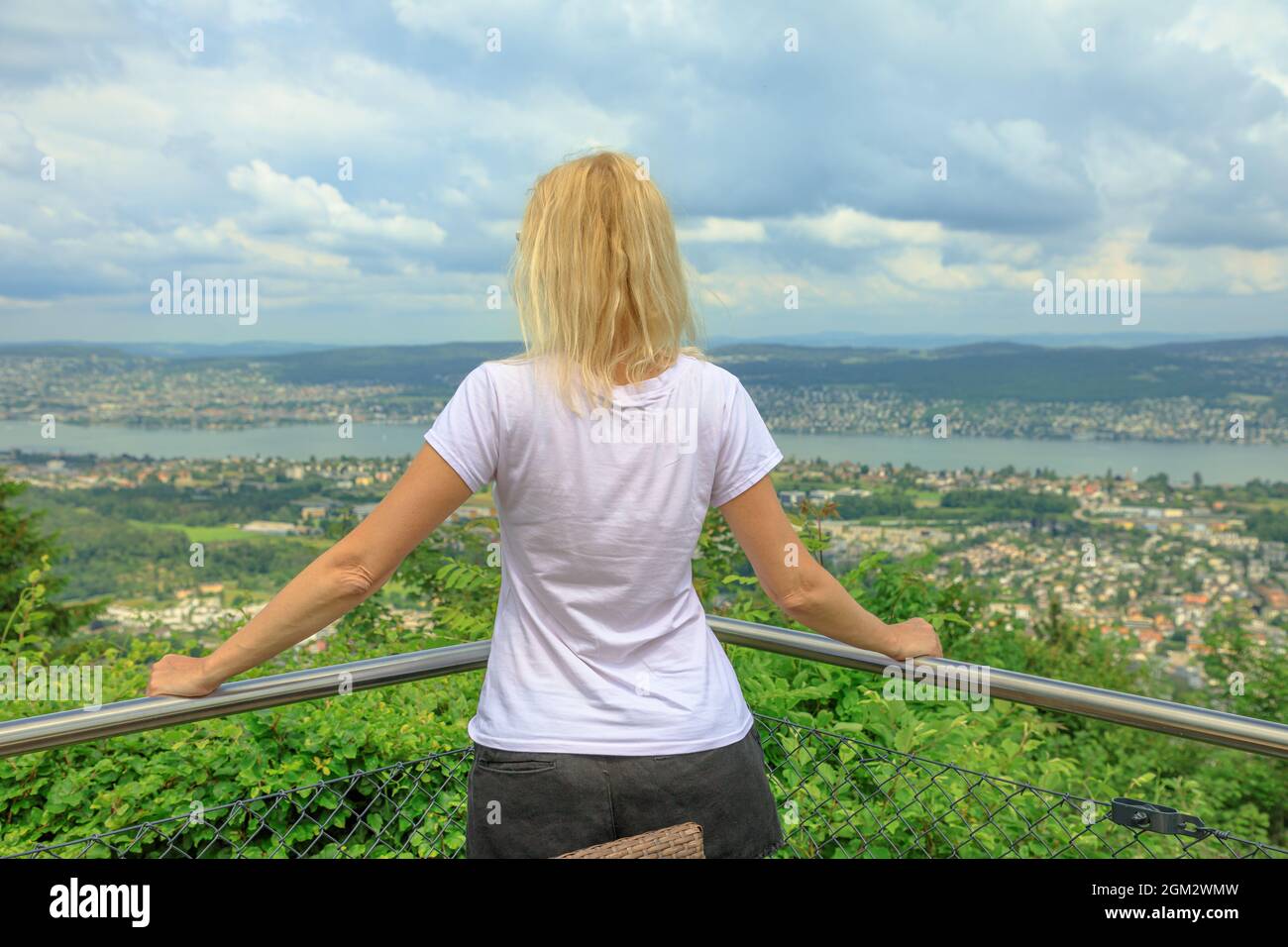 backside of woman looking Zurich city lake and skyline from top of Felsenegg cable car. Blonde tourist woman by lakefront in Deutch Switzerland of Stock Photo