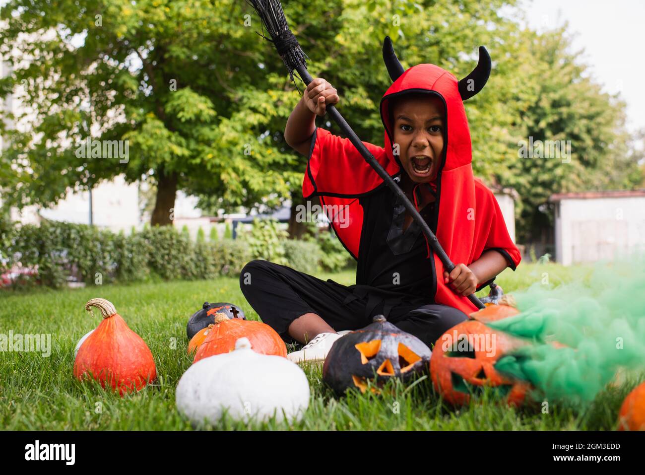 scary african american kid in devil halloween costume screaming while holding broom near pumpkins and sitting on lawn Stock Photo