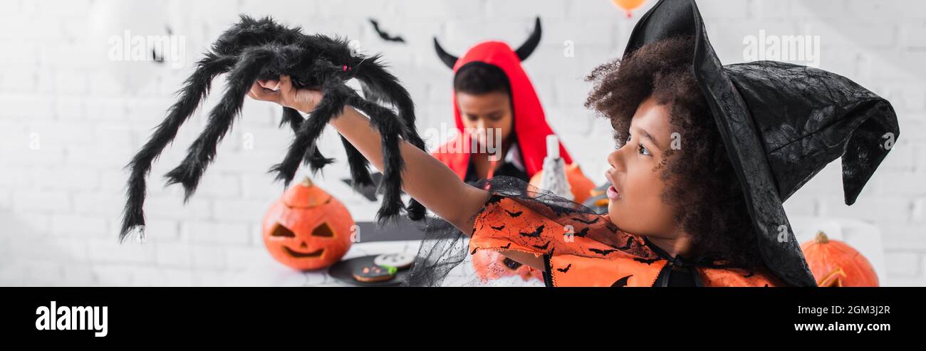 african american girl in witch costume holding toy spider near blurred brother, banner Stock Photo
