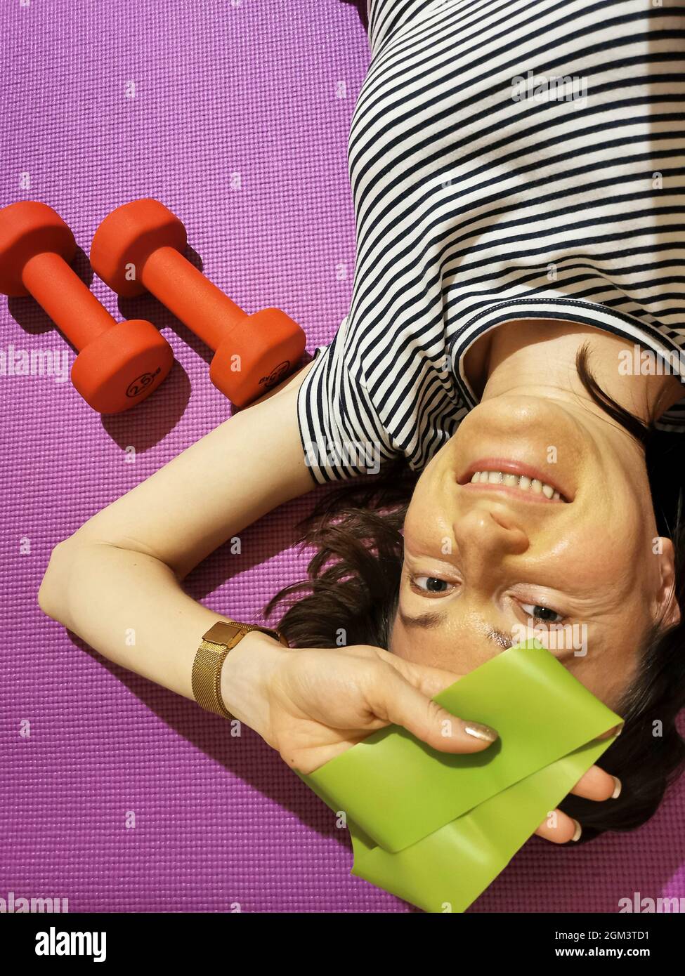 A smiling girl, tired of exercising, is lying on a yoga mat with 2.5 kg weights Stock Photo