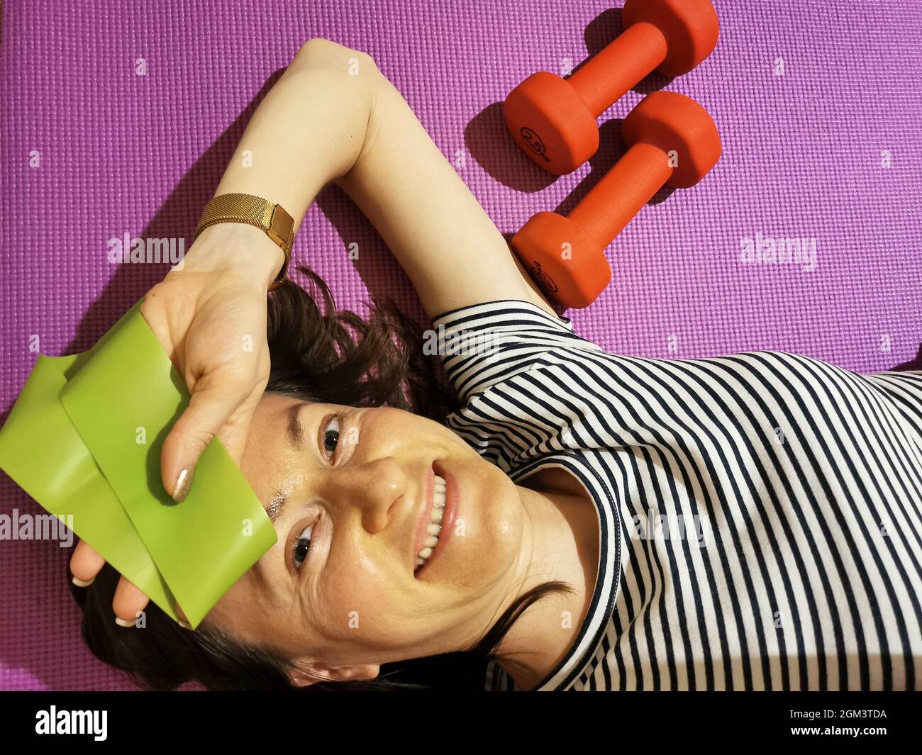 A smiling girl, tired of exercising, is lying on a yoga mat with 2.5 kg weights Stock Photo