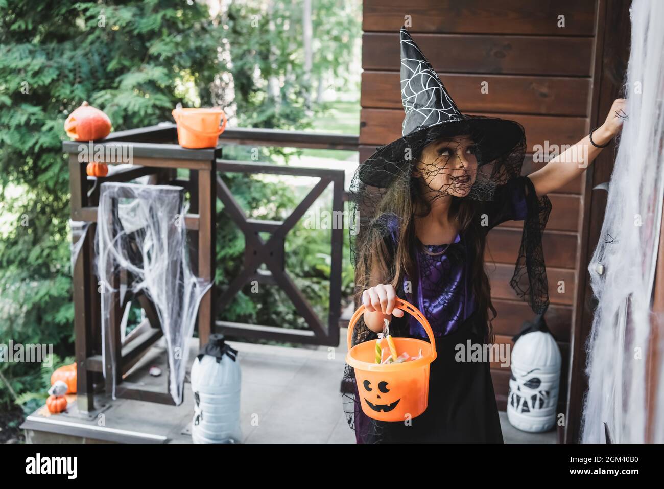 child in witch halloween costume holding bucket with sweets while knocking at door on decorated porch Stock Photo