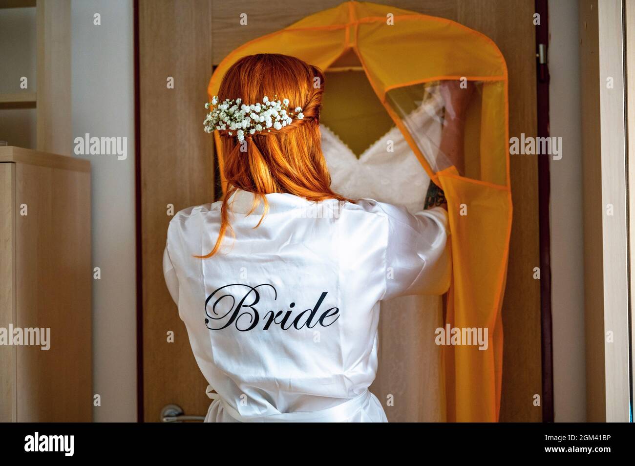 Red-haired women with wreath of flowers in white satin robe with inscription 'bride' takes off wedding dress from hanger. Bride preparing on wedding d Stock Photo