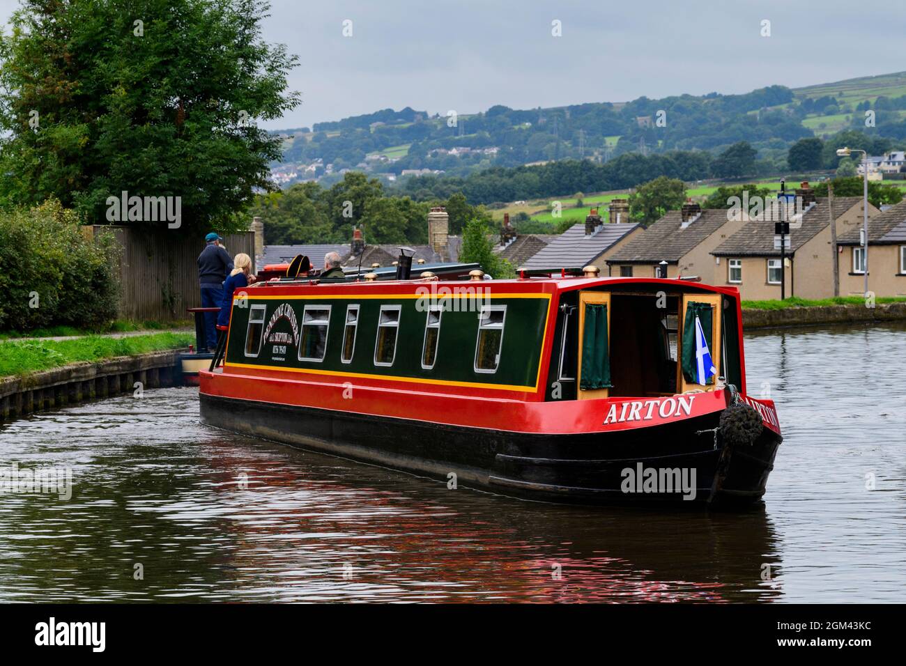 Hired red & green narrowboat sailing & travelling (casting-off) on semi-urban waterway, couple aboard - Leeds Liverpool Canal, Bingley, England, UK. Stock Photo