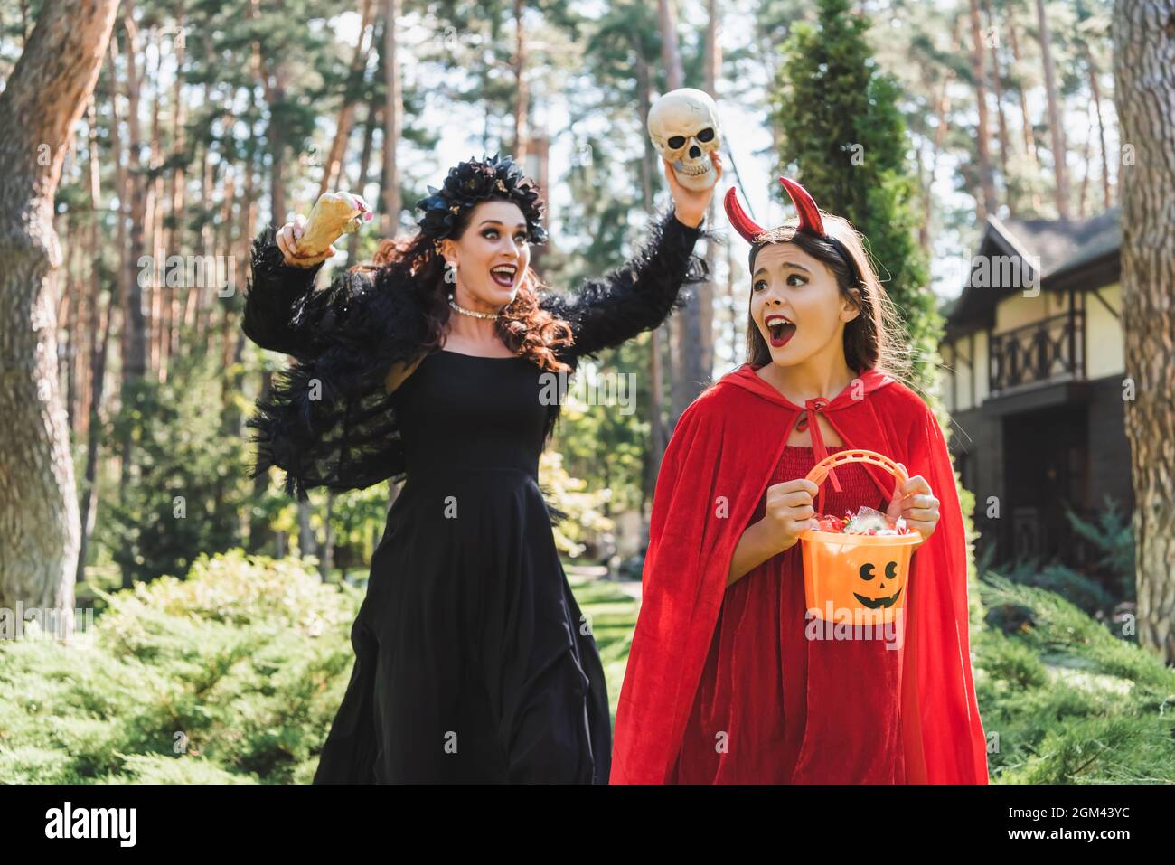 girl in devil halloween costume holding bucket with candies and screaming near mom with skull and toy hand Stock Photo