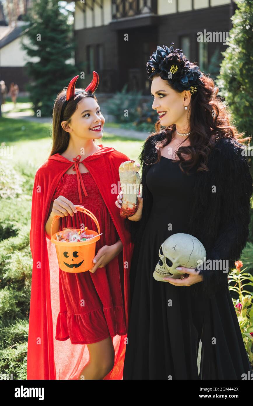 happy mom and daughter in vampire and devil halloween costumes, with bucket  of sweets, skull and toy hand in backyard Stock Photo - Alamy