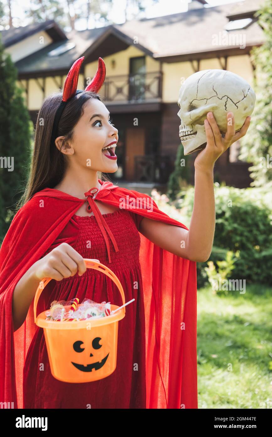 amazed girl in devil halloween costume looking at spooky skull while holding bucket with candies Stock Photo