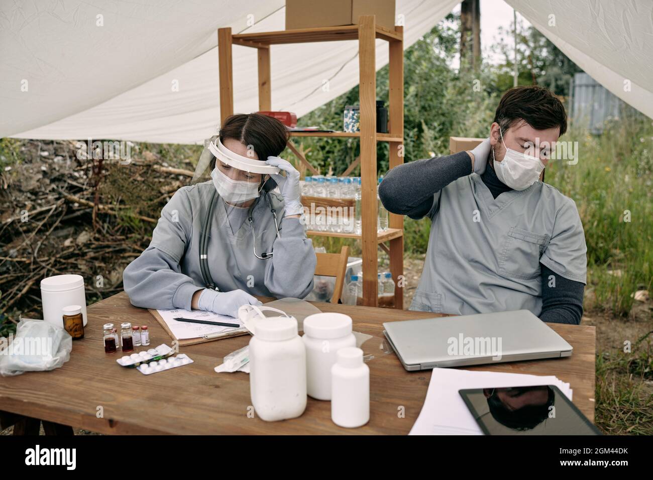 Exhausted ealthcare workers in masks sitting at table with pills under tent roof while working with refugees in camp Stock Photo
