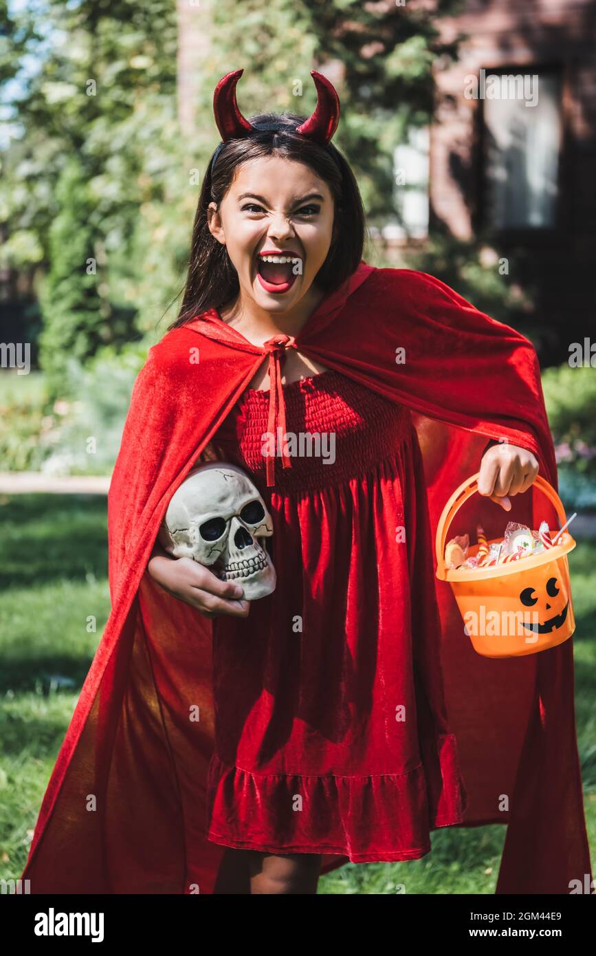 thrilled girl in devil halloween costume screaming while holding skull and bucket of candies Stock Photo