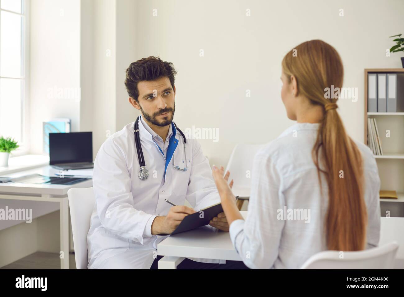 Attentive male doctor in hospital office consults female patient and records health complaints